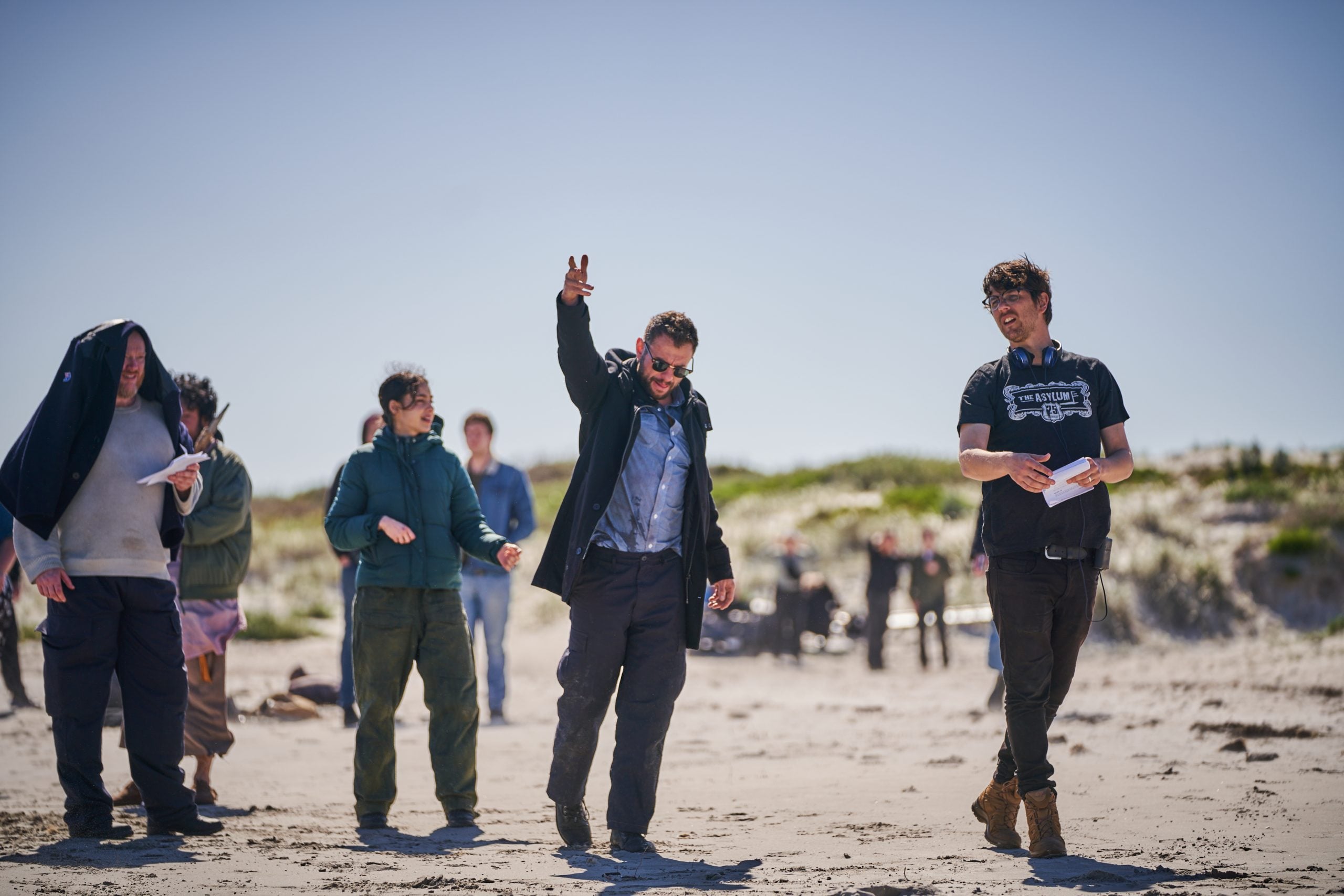 A group of people stand and walk on a sandy beach, some holding papers, with grassy dunes and a clear blue sky in the background.