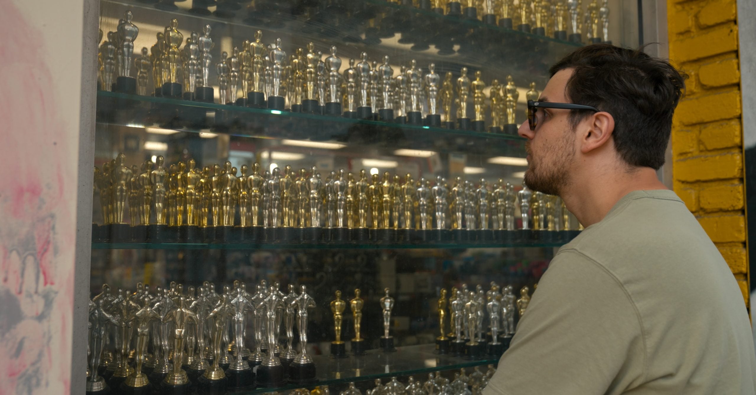 A man in sunglasses looks at shelves filled with gold and silver trophy statues inside a store display window.