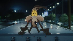 Animated Wile E. Coyote clings to the top of a moving truck at night, looking startled, with city streetlights blurred in the background.