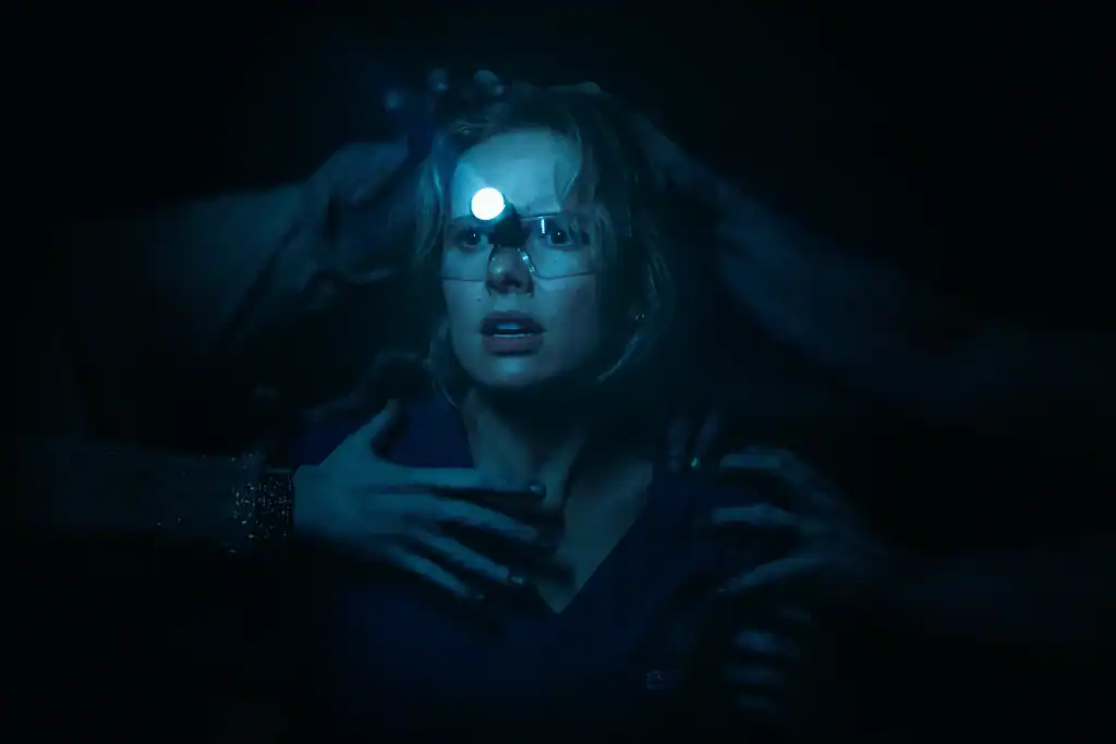 A woman wearing glasses and a headlamp looks startled as multiple hands reach toward her in a dark, eerie setting.