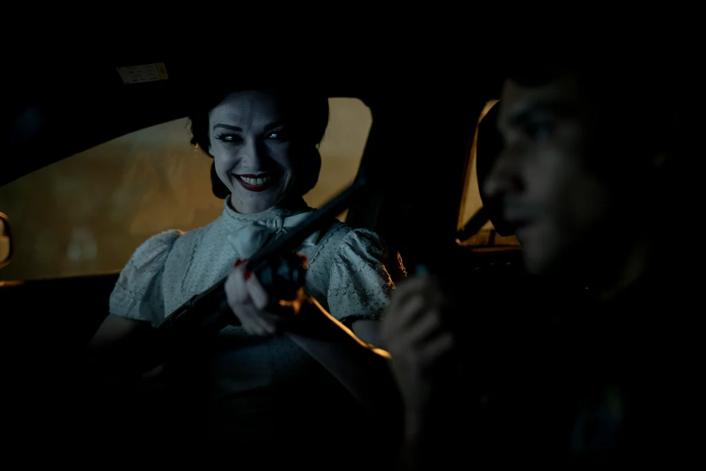 A woman in vintage clothing holds a shotgun and smiles menacingly at a man in a dark car; the man appears tense and speaks into a radio.