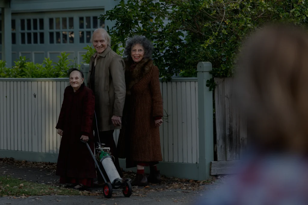 Three adults in brown coats stand by a fence; one holds a walker with an oxygen tank. Another person is blurred in the foreground.