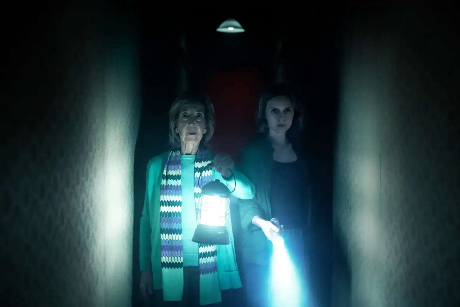 Two women stand in a dark hallway, one holding a lantern and the other shining a flashlight, both looking ahead with serious expressions.