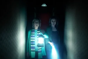 Two women stand in a dark hallway, one holding a lantern and the other shining a flashlight, both looking ahead with serious expressions.