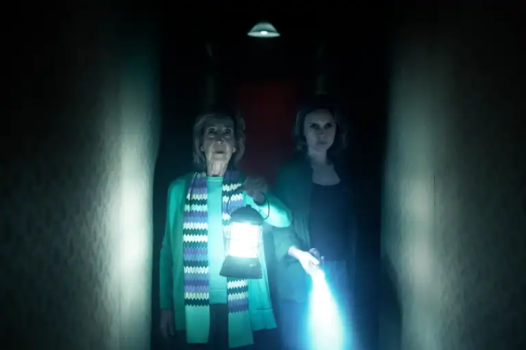 Two women stand in a dark hallway, one holding a lantern and the other shining a flashlight, both looking ahead with serious expressions.