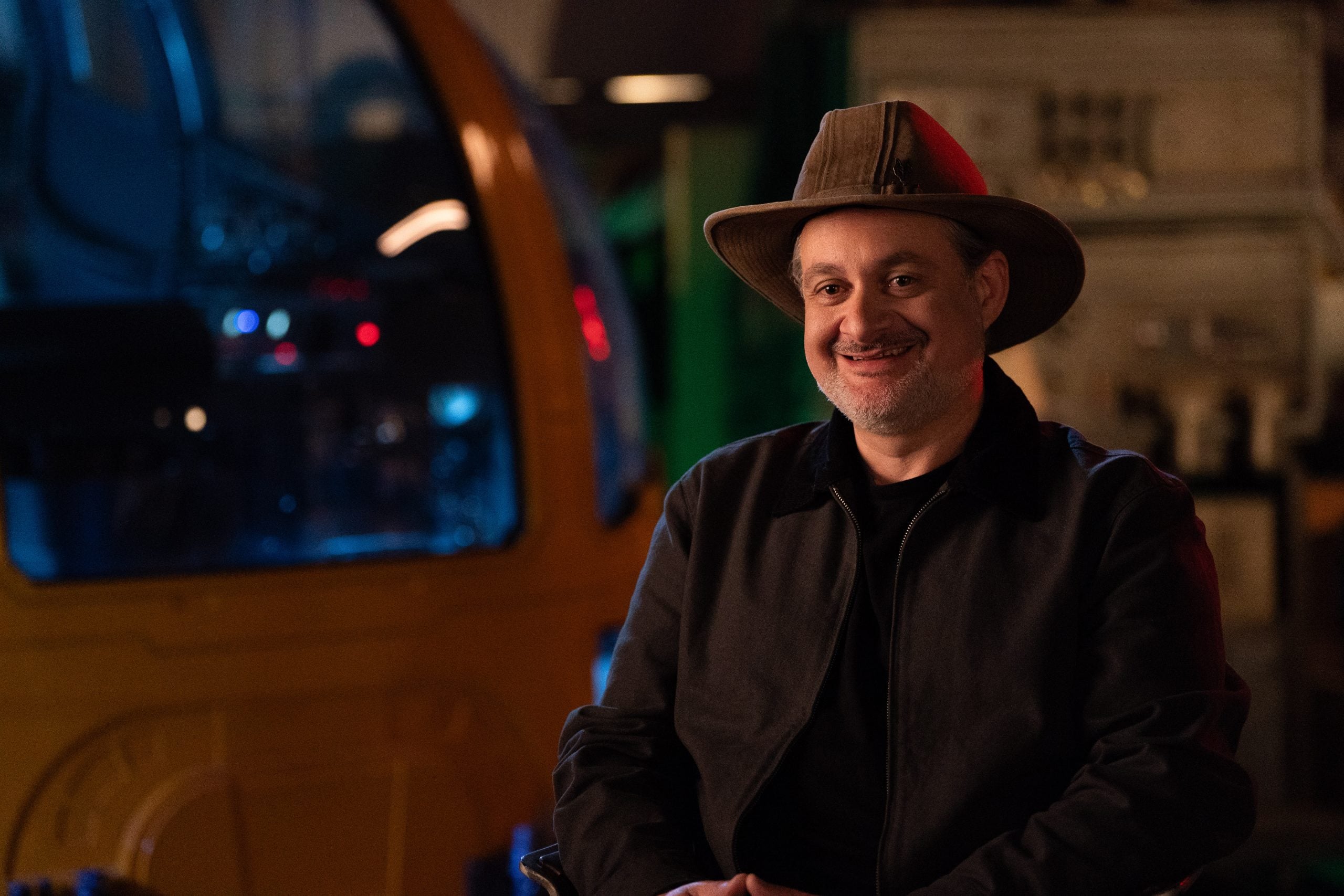 A man wearing a brown hat and dark jacket smiles while sitting indoors, with machinery and dim lighting in the background.
