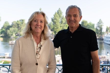 Two adults stand side by side outdoors near a body of water, with trees and a boat in the background. One is wearing a cream jacket, the other a black polo shirt.
