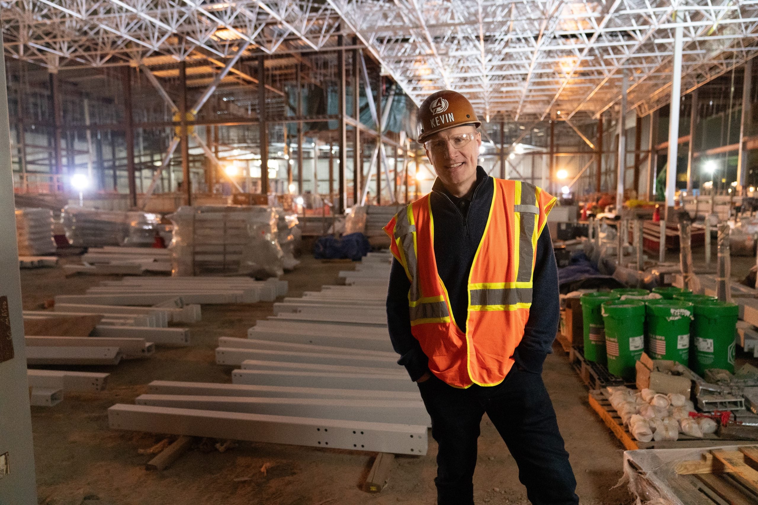 Person wearing a hard hat and reflective vest stands in a large, partially constructed industrial building with steel beams and construction materials in the background.