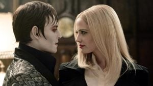 A pale man with slicked dark hair and a woman with long blonde hair face each other closely in a dimly lit room.