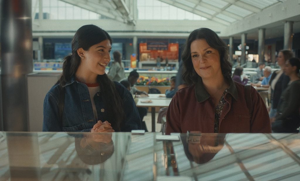 Two women stand side by side at a counter in a brightly lit mall food court, smiling and engaging in conversation.