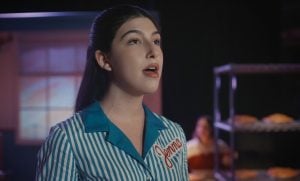 A woman in a striped uniform with “Jenna” embroidered sings on stage, with a rack of pies and another person blurred in the background.