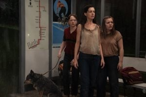 ‘The End Of Oak Street’ Review – Adrenaline Fueled Blockbuster Is The Best Dinosaurs Have Been On Screen In Decades Three people and a dog stand at a bus stop in the rain, looking concerned. A transit map and a backpack are visible on the bench beside them.