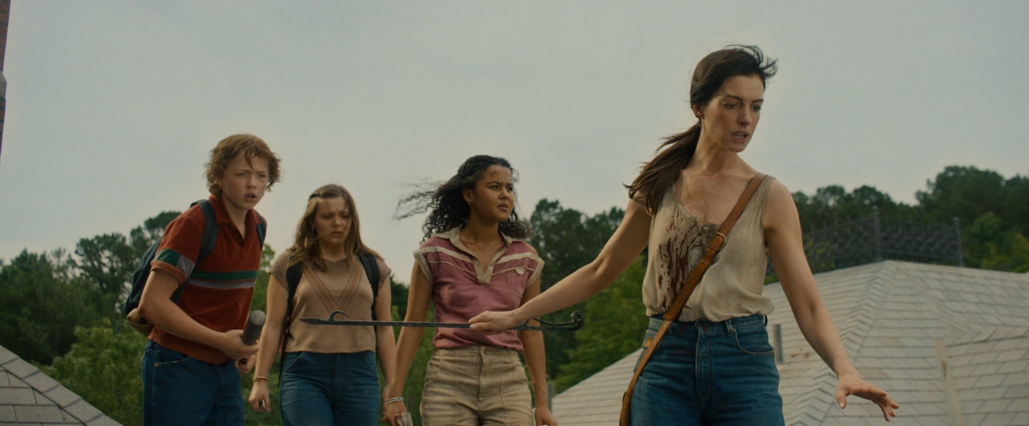 Four people stand on a rooftop looking alert; one woman in front holds a metal rod while the others watch, with trees and rooftops visible in the background.