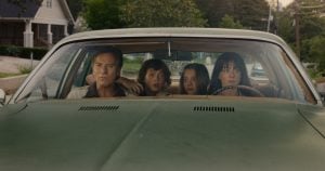 Four people sit in a green car, looking out the windshield with serious expressions; trees and houses are visible outside.