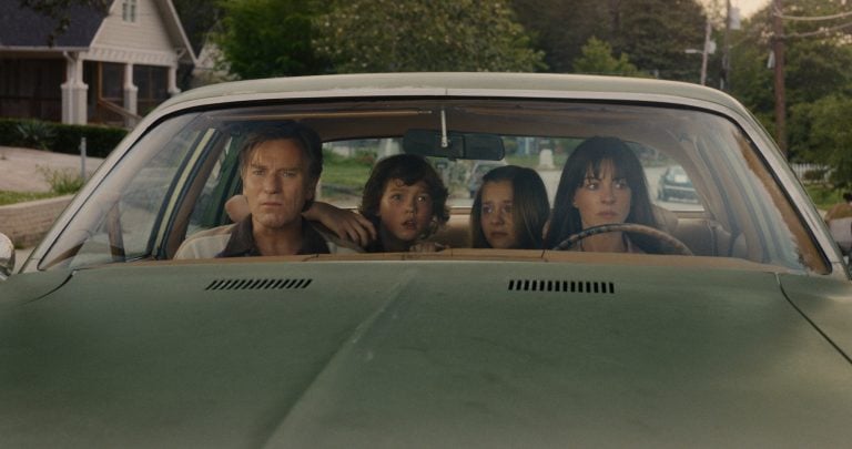 Four people sit in a green car, looking out the windshield with serious expressions; trees and houses are visible outside.