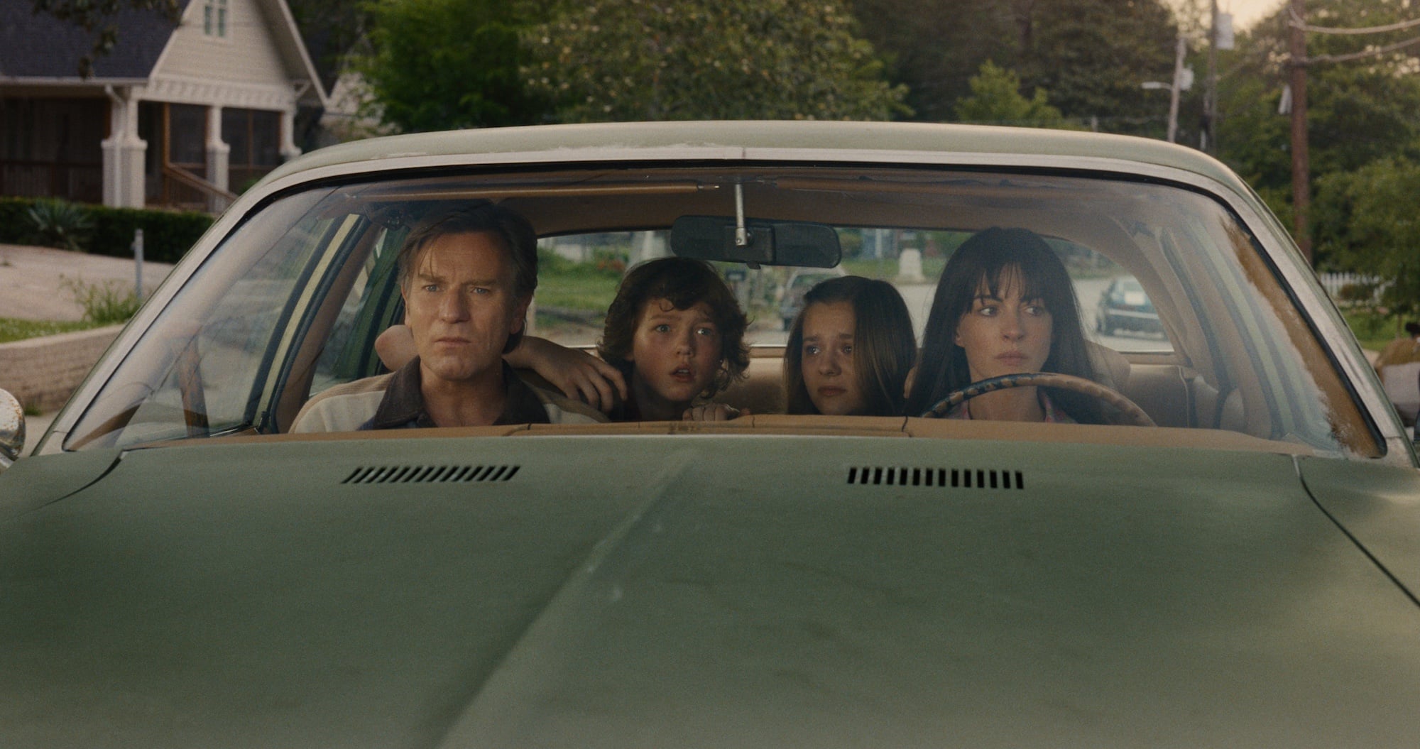 Four people sit in a green car, looking out the windshield with serious expressions; trees and houses are visible outside.