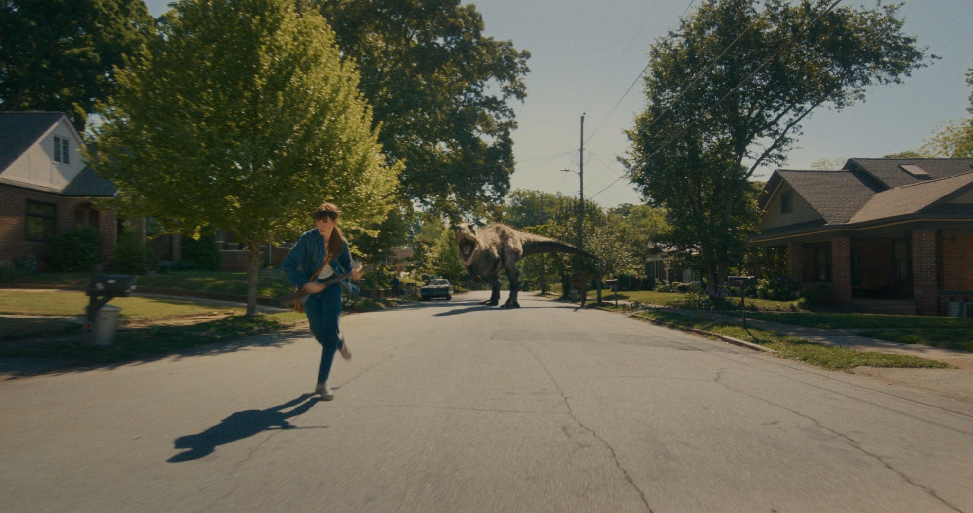 A person runs down a suburban street while being chased by a large dinosaur in broad daylight.