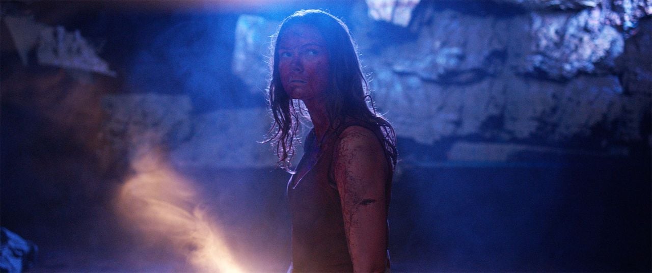 A woman with disheveled hair and a dirty face stands in a dim, rocky cave illuminated by blue and orange light.