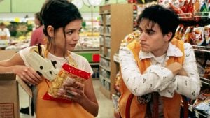 Two grocery store workers in orange vests talk in an aisle; one holds a pricing gun and a bag of chips, the other leans on a shelf.