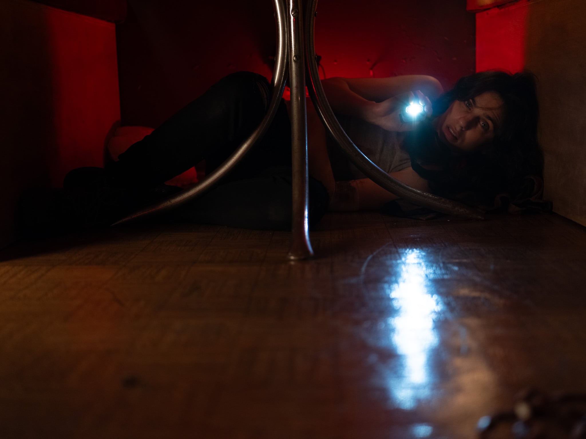 A person lies on the floor under a table, holding a flashlight with a beam reflecting on the floor. The scene is dimly lit with red light in the background.