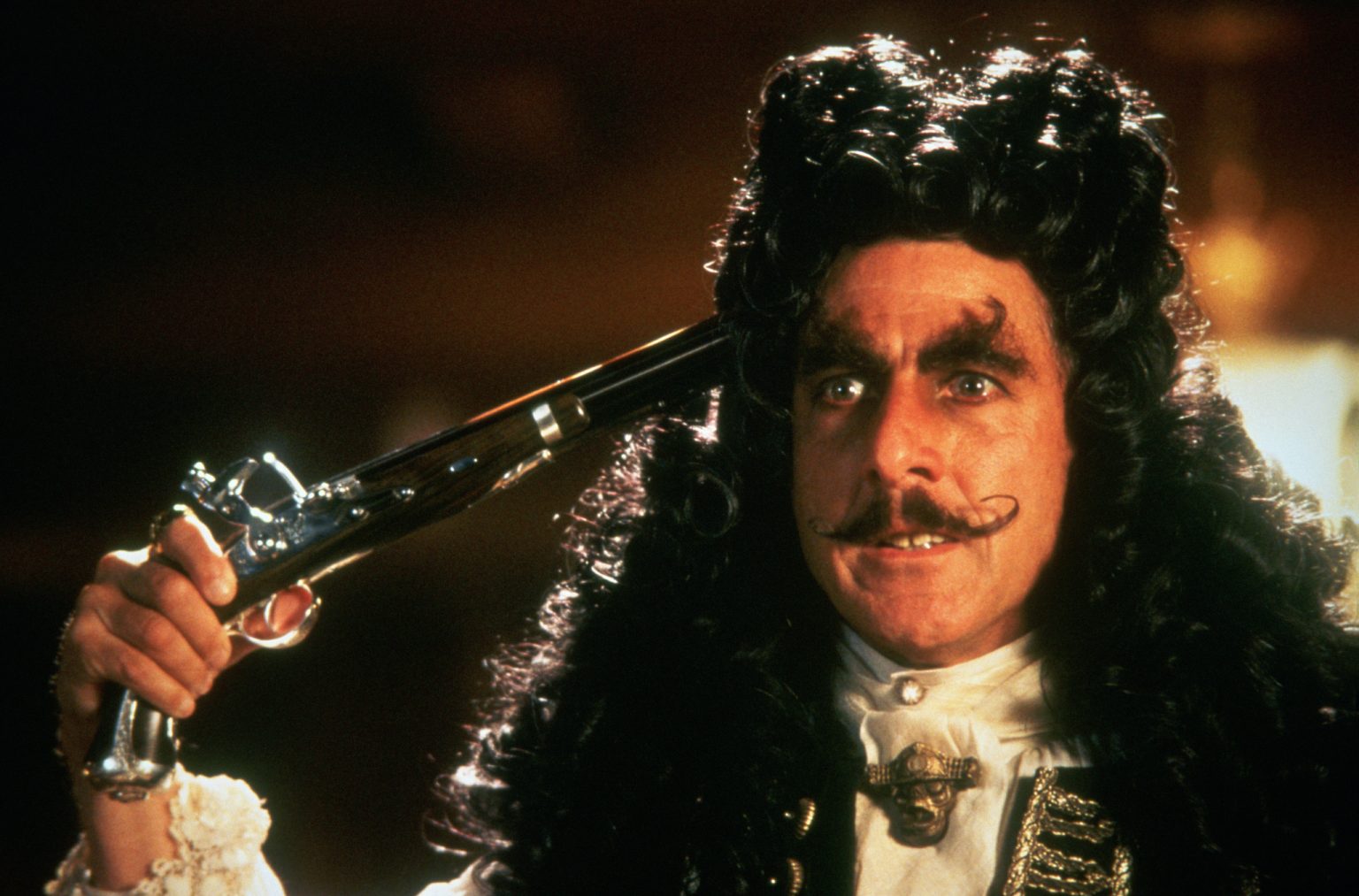 A man in elaborate 18th-century costume and wig holds a vintage pistol to his head, with a dramatic expression and curled mustache.