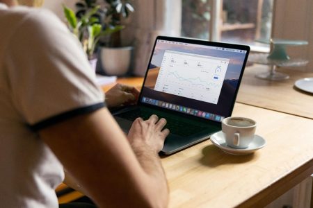 Person working on a laptop displaying analytics charts at a wooden table with a cup of coffee and potted plants nearby.