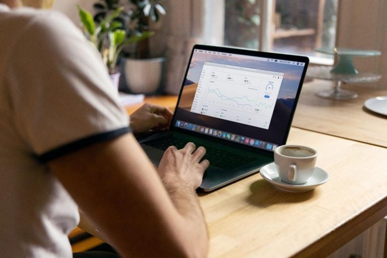 Person working on a laptop displaying analytics charts at a wooden table with a cup of coffee and potted plants nearby.