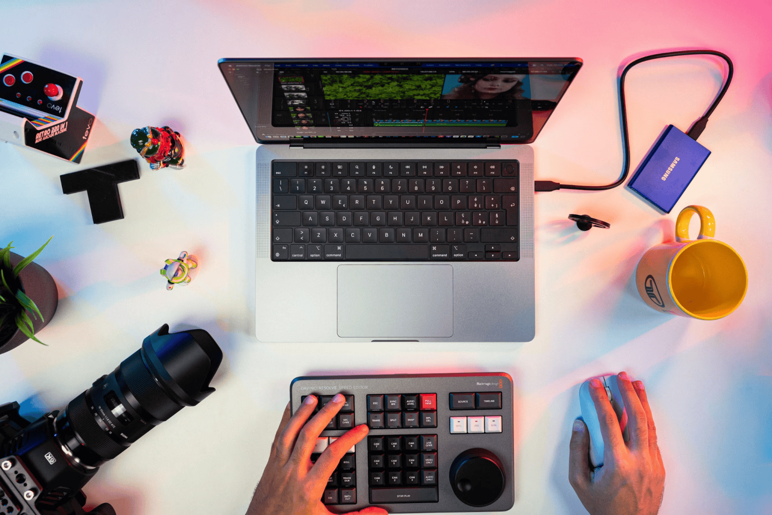 Overhead view of hands using a laptop and control panel at a desk with a camera, external drive, mug, plant, and small figurines.