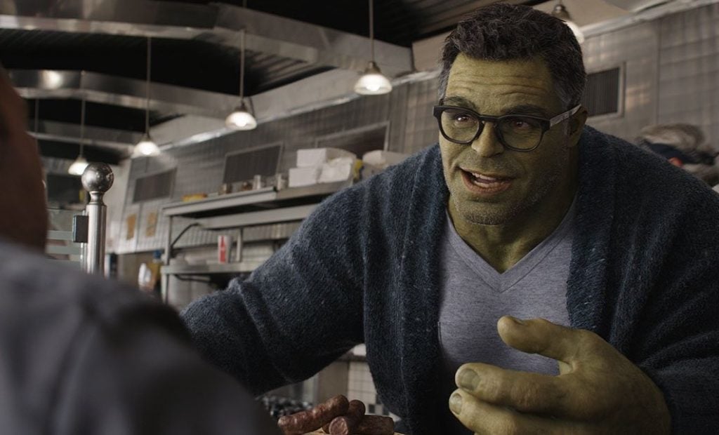 A large green humanoid with glasses and a sweater speaks to someone across a diner counter.