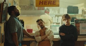 Three men stand at a counter beneath a yellow “HERE” sign, with a worker visible preparing food in the background of a casual restaurant.