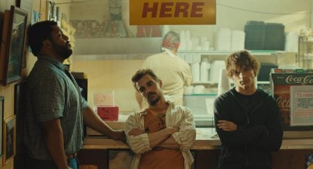 Three men stand at a counter beneath a yellow “HERE” sign, with a worker visible preparing food in the background of a casual restaurant.