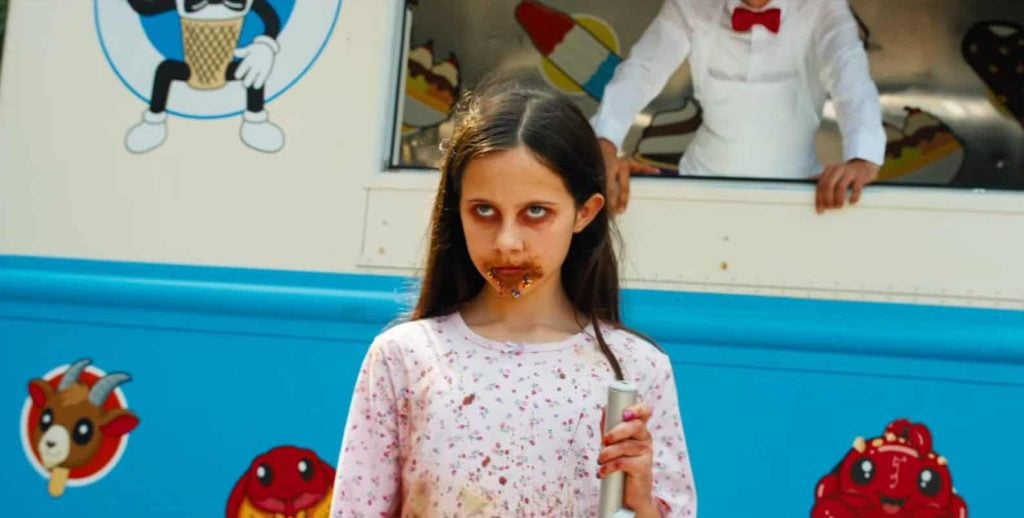 A girl with messy face and dark eye makeup stands in front of an ice cream truck, holding a metal object. An adult in a white uniform is visible in the truck window.
