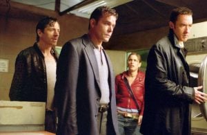 Four people stand in a dimly lit laundry room, looking tense and serious; one person is near a washing machine.