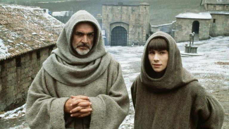 Two people in brown hooded robes stand in a medieval courtyard with stone buildings and a statue in the background on a cold day.