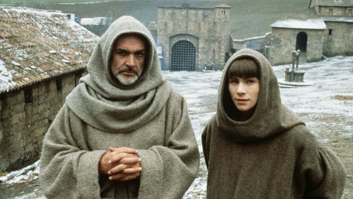 Two people in brown hooded robes stand in a medieval courtyard with stone buildings and a statue in the background on a cold day.