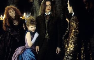Four people in elaborate, old-fashioned costumes stand indoors; three appear tense while one woman in blue clings to the man in the center.
