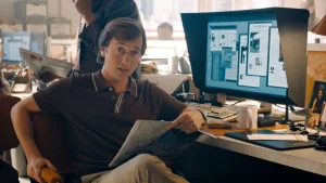 ‘The People V. Gorilla Grodd’ Is Official As HBO Max Orders Eight-Episode DCU Series A man sits at a cluttered office desk holding a newspaper, with a large computer monitor displaying page layouts in the background.