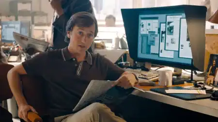 A man sits at a cluttered office desk holding a newspaper, with a large computer monitor displaying page layouts in the background.