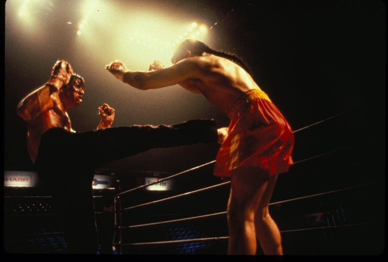 Two martial artists spar in a boxing ring under bright arena lights, one delivers a kick to the other’s chest while both wear shorts and are shirtless.