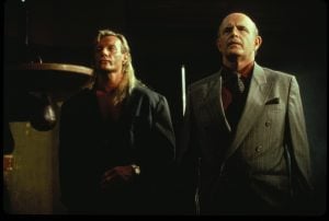 Two men stand indoors; one has long blonde hair and wears a dark open shirt, the other is bald, wearing a gray pinstripe suit and tie.