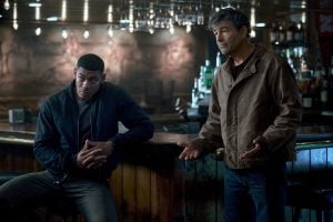 Two men stand in a dimly lit bar; one leans against the counter with hands clasped, while the other gestures with open hands, both appearing serious.
