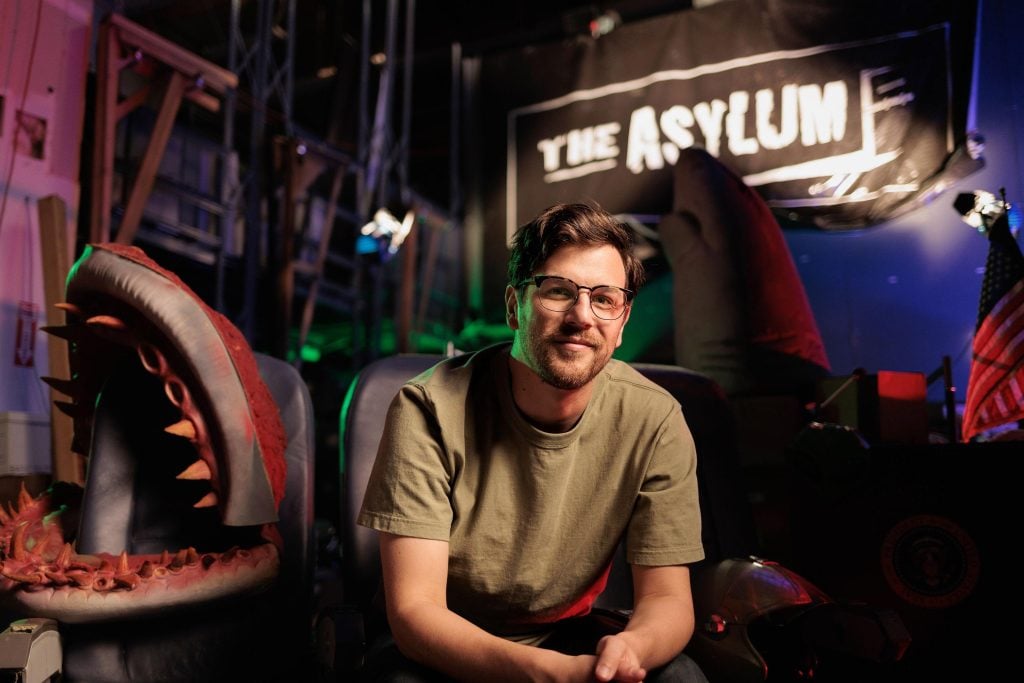 A man with glasses sits between two large shark props in a dimly lit room with a sign reading “The Asylum” in the background.