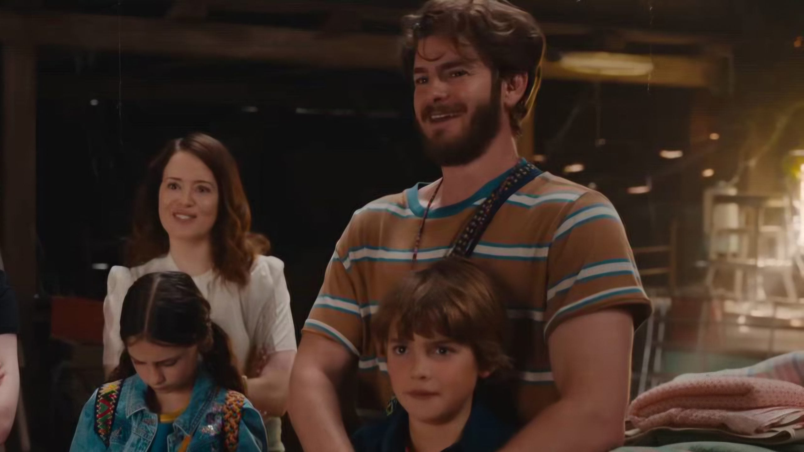 A man smiles while standing with a woman and two children indoors; one child looks down, the other faces forward.