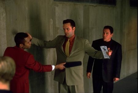 Lionsgate To Release The Jon Favreau & Vince Vaughn Crime Comedy ‘Made’ On 4K UHD Blu-Ray A man in a suit holds up an ID while being frisked with a handheld scanner by security, as another man waits nearby against a concrete wall.