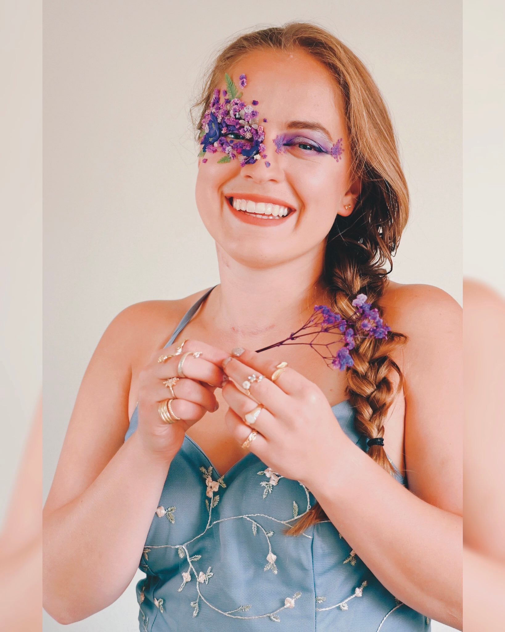 Woman in a blue dress with embroidered flowers, smiling, holding purple dried flowers, with purple and pink floral makeup covering one eye.