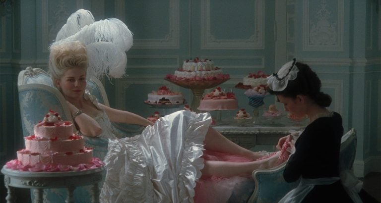 A woman in an elaborate white dress lounges on a blue chair, surrounded by cakes, while a maid tends to her feet in an ornate pastel room.
