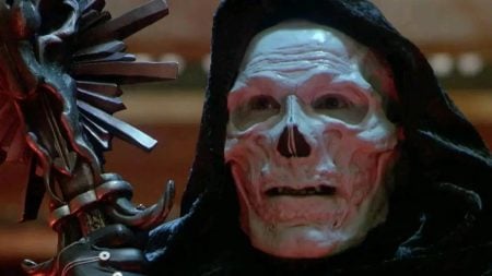 A person in a hooded cloak wears a white skull mask and holds a staff with metallic, spiked details.