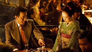 A man in a suit gestures while seated at a table with a woman in a traditional kimono in a dimly lit restaurant filled with people.