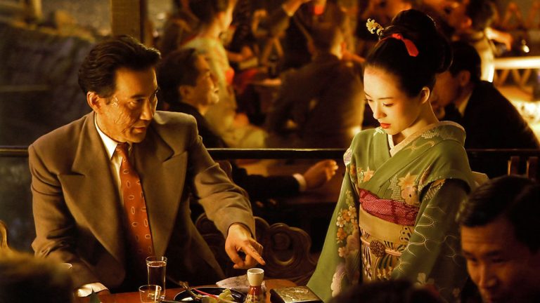 Sony To Release The Epic Period Drama ‘Memoirs Of A Geisha’ On 4K UHD Blu-Ray This November A man in a suit gestures while seated at a table with a woman in a traditional kimono in a dimly lit restaurant filled with people.