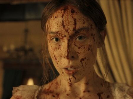 "A young woman with long hair, dressed in light clothing, has her face and upper chest covered in blood, standing indoors with dim warm lighting and blurred curtains and furnishings in the background."